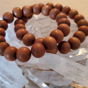 May include: Two wooden bead bracelets. The beads are a warm brown color and appear to be made of wood. The bracelets are resting on a bed of clear, crystalline quartz.