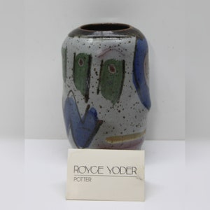 May include: A ceramic vase with a speckled gray background and abstract blue, green, and purple painted shapes. The vase has a dark brown rim and a label that reads "ROYCE YODER POTTER".