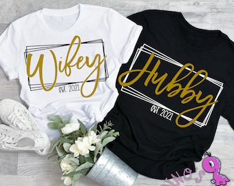 May include: Two t-shirts with a gold script design. The white t-shirt says "Wife" with a black and white frame and the text "Est. 2021". The black t-shirt says "Hubby" with a white and black frame and the text "Est. 2021".