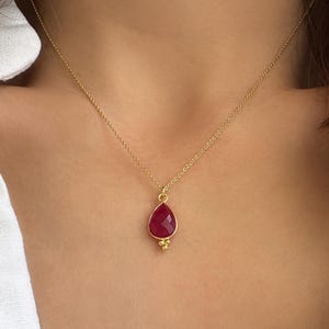 May include: A gold chain necklace featuring a faceted, teardrop-shaped dark pink gemstone pendant. The pendant is set in a gold bezel with three small gold spheres at the bottom point, suspended from a delicate gold link chain that rests on the neck.