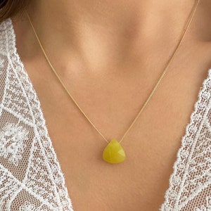 Puede incluir: Un delicado collar de oro con un colgante triangular de piedra preciosa amarilla. El colgante cuelga de una fina cadena de oro. El collar se lleva sobre un top de encaje blanco.