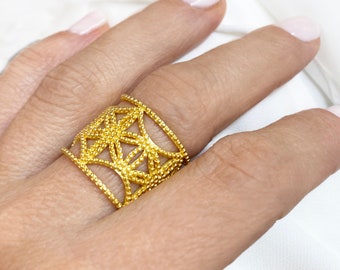 May include: A gold-colored ring with an openwork design featuring geometric star-like patterns. The ring is worn on a finger, showcasing its intricate details and bright, shiny finish. The ring's design is made of small, round beads.