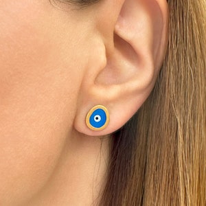 Puede incluir: Un par de pendientes de color dorado con un diseño de ojo turco azul. Los pendientes son pequeños y delicados, y serían un gran regalo para alguien especial.