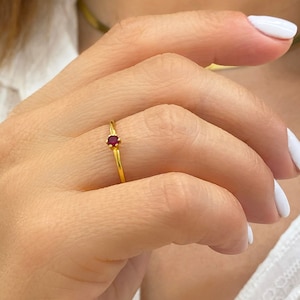 May include: A slender gold ring with a small, round, red gemstone. The ring is set on a thin gold band. The image shows the ring on a finger, showcasing the jewellery's understated elegance.