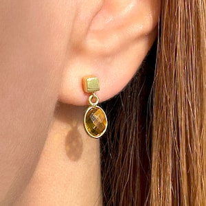 Puede incluir: Pendientes de oro con un pequeño dije cuadrado y una piedra preciosa ovalada colgante. La piedra preciosa es de color ámbar oscuro.