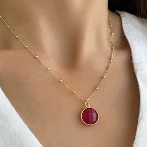 Op de afbeelding: Een goudkleurige ketting met een fijne schakelketting voorzien van regelmatig geplaatste kleine gouden kraaltjes. De ketting heeft een ronde hanger met een gefacetteerde, diep magentakleurige edelsteen in een goudkleurige metalen zetting.