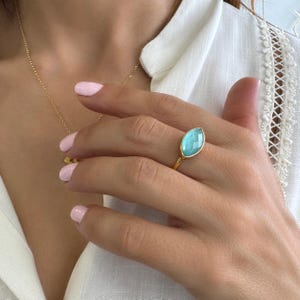 Peut inclure: Une bague en or ornée d'une pierre précieuse bleu clair de forme marquise. La bague est sertie dans un anneau en or et portée au doigt. Un collier en or est également visible. L'arrière-plan comprend une chemise blanche.