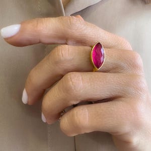 Puede incluir: Un anillo de oro con una gema rosa intenso en forma de marquesa. El engaste del anillo es una banda de oro pulido. La gema facetada refleja la luz, mostrando su color vibrante. El anillo se muestra en una mano con esmalte de uñas blanco.