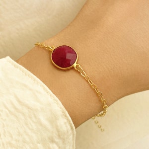 Könnte beinhalten: Ein goldfarbenes Armband mit einem großen, oval geschliffenen, facettierten Rubin. Der Rubin ist in einer goldenen Lünette gefasst und mit einer feinen Kette verbunden. Das Armband wird am Handgelenk getragen, mit einem weißen Hemdsärmel im Hintergrund.