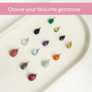 Puede incluir: Una colecci&oacute;n de gemas en forma de l&aacute;grima de varios colores, incluyendo verde, azul y morado, dispuestas sobre una bandeja blanca. Cada gema tiene un peque&ntilde;o lazo de metal. El texto "Choose your favourite gemstone" aparece en la parte superior.