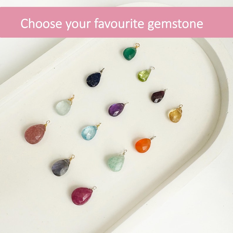 Puede incluir: Una colecci&oacute;n de gemas en forma de l&aacute;grima de varios colores, incluyendo verde, azul y morado, dispuestas sobre una bandeja blanca. Cada gema tiene un peque&ntilde;o lazo de metal. El texto "Choose your favourite gemstone" aparece en la parte superior.