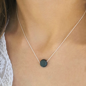 Puede incluir: Un collar de plata con un colgante circular gris oscuro. El colgante cuelga de una delicada cadena de plata. El collar se muestra en el pecho de una persona.