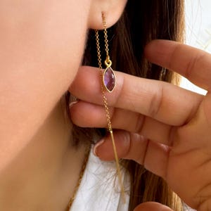 Puede incluir: Pendientes colgantes dorados con una gema púrpura en forma de marquesa colgando. El pendiente se muestra en uso, con la cadena extendiéndose hacia abajo. La gema está engastada en un bisel dorado.