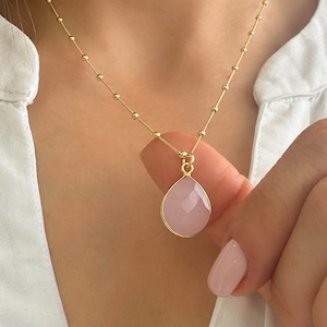 Pode incluir: Um colar dourado com uma corrente delicada com pequenas contas espaçadas uniformemente. O pingente é uma pedra preciosa rosa facetada em forma de gota, emoldurada em uma moldura dourada. O colar está sendo segurado por uma pessoa.