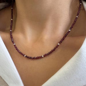 Op de afbeelding: Een delicate ketting met kleine, ronde, dieprode kralen afgewisseld met kleine zilverkleurige kralen. De ketting wordt om de hals gedragen en toont zijn eenvoudige elegantie en subtiele glans. De kralen zijn gelijkmatig verdeeld, wat een verfijnde en klassieke uitstraling creëert.