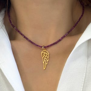 Könnte beinhalten: Eine goldene Halskette mit einem zarten Flügel-Anhänger hängt an einem Modellhals. Die Halskette besteht aus kleinen, violetten Perlen.