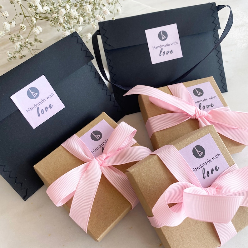 Puede incluir: Vista a&eacute;rea de cajas de regalo y sobres negros. Las cajas marrones est&aacute;n atadas con cintas rosas. Cada caja y sobre tiene una pegatina que dice "Handmade with love".