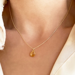 May include: A delicate gold necklace featuring a small, teardrop-shaped pendant. The pendant has a warm, amber hue and is suspended from a fine gold chain. The necklace is shown against a neutral background.
