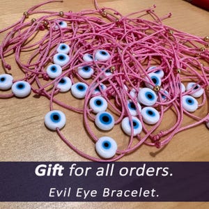 Puede incluir: Una colecci&oacute;n de pulseras de cord&oacute;n rosa, cada una con un dije de ojo turco blanco y azul. Las pulseras tienen cuentas doradas y est&aacute;n dispuestas sobre una superficie de madera. El texto "Evil Eye Bracelet" est&aacute; incluido.