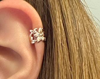 Puede incluir: Un pendiente tipo ear cuff de oro rosa con un diseño floral detallado que presenta una flor central de cinco pétalos rodeada por motivos delicados de enredaderas y hojas. El accesorio metálico se sitúa en el borde exterior del cartílago.