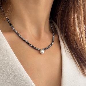 Può includere: Una collana di perline grigio scuro con un singolo pendente rotondo color argento. La collana è indossata su un top bianco, creando un contrasto. Le perline sembrano piccole e uniformemente distanziate.