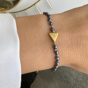 Könnte beinhalten: Ein Armband aus schwarzen Hämatitperlen mit einem goldenen Dreiecksanhänger.