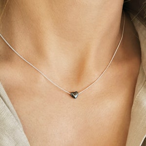 Könnte beinhalten: Eine zarte silberfarbene Halskette mit einem einzelnen, kleinen, polierten, dunkelgrauen herzförmigen Anhänger, der mittig an der Kette sitzt. Die Kette liegt am Hals an und zeigt ein minimalistisches Design mit glatter, reflektierender Oberfläche.