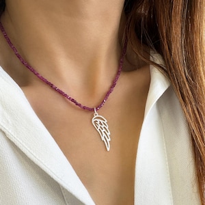 Könnte beinhalten: Eine Halskette mit einem silbernen Engelsflügel-Anhänger und einer Kette aus violetten Edelsteinperlen.