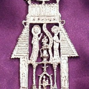 Op de afbeelding: Een zilveren hanger die een religieuze scène voorstelt met drie figuren in een gebouw. De hanger is gedetailleerd met ingewikkelde houtsnijwerk en een kruis aan de bovenkant.