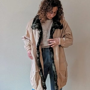 Può includere: Una donna indossa una lunga giacca beige con un colletto di pelliccia nera, jeans blu e scarpe da ginnastica blu chiaro.