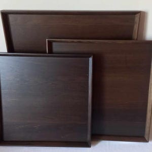Könnte beinhalten: Drei dunkelbraune Holztabletts in verschiedenen Größen sind gestapelt. Die Tabletts haben eine rechteckige Form mit erhöhten Kanten. Die Holzmaserung ist sichtbar und die Oberfläche ist glatt. Die Tabletts scheinen neu zu sein.
