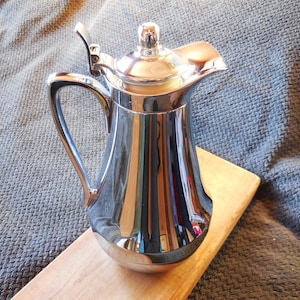 May include: A polished silver jug with a curved handle and a hinged lid. The jug is placed on a light-coloured wooden board, against a grey textured backdrop. The jug's surface reflects the surrounding environment.
