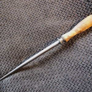 May include: A sharpening steel featuring a long, tapered, metallic rod with a smooth surface and a rounded tip. The tool includes a small, circular metal guard and a light-coloured, weathered wooden handle with a visible grain and worn texture.