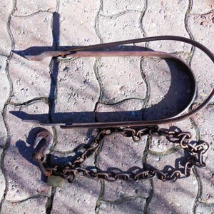 May include: A rusty metal chain and hook attached to a metal loop. The chain and loop are lying on a gray concrete surface.