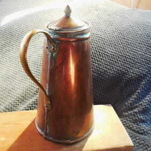 May include: A copper teapot with a brass handle and a lid with a finial. The teapot has a patina and is sitting on a wooden surface.