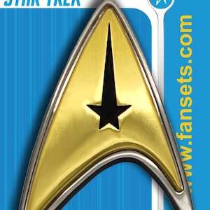 May include: A gold and black Star Trek communicator badge. The badge is delta-shaped with a black star in the centre. The words "STAR TREK" are at the top, and "ADULT COLLECTIBLE" is at the bottom.