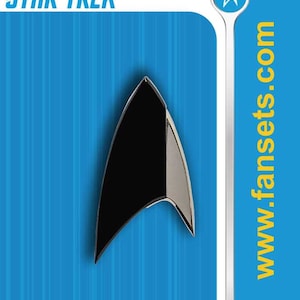 May include: A Star Trek delta-shaped pin with a black center and silver metallic border, mounted on a blue display card featuring the text STAR TREK, www.fansets.com, and ADULT COLLECTIBLE. The pin has a pointed top and curved side edges.
