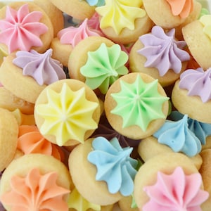 Puede incluir: Primer plano de una pila de galletas pequeñas y redondas. Cada galleta está cubierta con un remolino de glaseado en colores pastel: rosa, morado, amarillo, verde, naranja y azul. Las galletas son de color marrón claro.