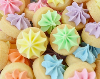 May include: A close-up shot of a pile of small, round cookies. Each cookie is topped with a swirl of pastel-colored frosting in shades of pink, purple, yellow, green, orange, and blue. The cookies are a light tan color.