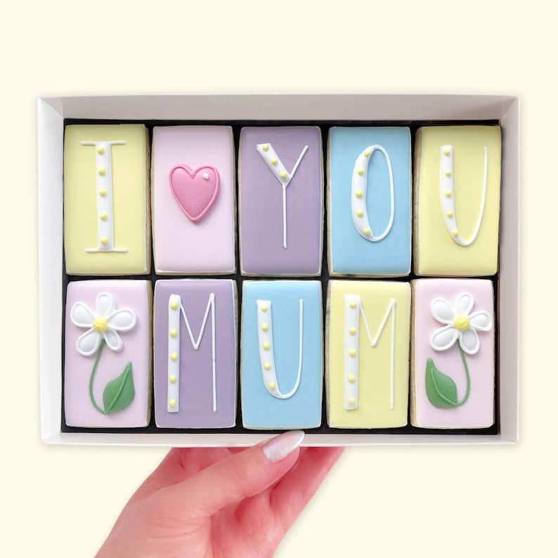 A selection of hand-iced biscuits in an I Love You Mum theme against a black background, held in a hand featuring ten blocks piped with I Love You Mum and two white flowers, decorated in pastel yellow, pink, purple and blue