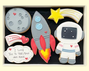 May include: A set of six decorated sugar cookies in a white box. The cookies are space-themed and include a rocket, an astronaut, a moon, a shooting star, and a cloud with the text "I love you to the moon and back".