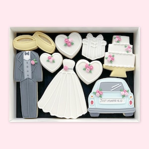 Puede incluir: Una caja de galletas decoradas con temática de boda. Las galletas incluyen un esmoquin, un vestido de novia, corazones con flores rosas, un regalo, una tarta de varios pisos, un coche con el texto "Just Married" y anillos de boda.