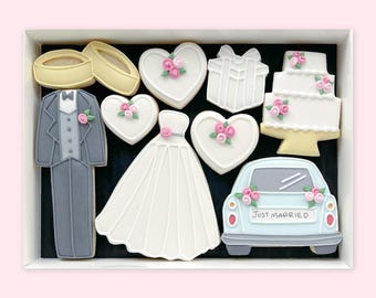 May include: A box of wedding-themed biscuits. The selection includes a tuxedo, wedding dress, heart-shaped biscuits with pink flowers, a gift, a tiered cake, a car with "Just Married" text, and wedding rings.