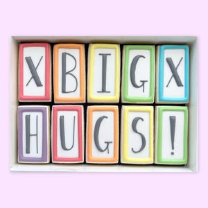 May include: A box of decorated biscuits spelling out "X BIG X HUGS!". Each biscuit is rectangular with a white background and a different coloured border. The letters are grey.