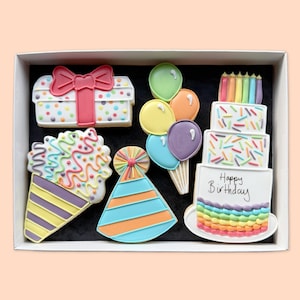 Puede incluir: Un conjunto de cinco galletas decoradas en una caja blanca. Las galletas incluyen una caja de regalo con un lazo rosa, un grupo de globos coloridos, un pastel de tres niveles con el texto Happy Birthday, un gorro de fiesta y un cono glaseado.