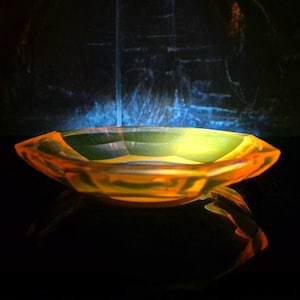 Könnte beinhalten: Eine flache, ovale Glasschale in Orange-Gelb mit facettiertem Design. Die Schale steht auf einer reflektierenden Oberfläche. Der dunkle Hintergrund weist blaue und weiße Akzente auf, die einen Kontrast zur Farbe der Schale bilden.
