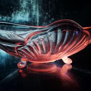 Könnte beinhalten: Eine Vintage-Glasschale in Rosa mit einem muschelartigen Design. Die Schale hat einen gewellten Rand und steht auf drei kleinen Füßen. Das Glas reflektiert das Licht und erzeugt einen Glanzeffekt. Die Schale wird wahrscheinlich zum Servieren oder Ausstellen verwendet.