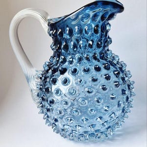 Könnte beinhalten: Eine blaue Glaskanne mit strukturiertem Hobnail-Design. Die Kanne hat einen klaren, gebogenen Griff und einen gerüschten Rand. Das Glas ist leuchtend blau und das Hobnail-Muster bedeckt die gesamte Oberfläche der Kanne.