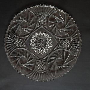 Puede incluir: Un plato para pastel de vidrio transparente con un patrón decorativo intrincado. El plato tiene un borde festoneado y un diseño central elevado.