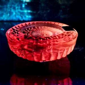 Könnte beinhalten: Ein roter, kreisförmiger Aschenbecher aus Glas mit strukturiertem Rand und einer kleinen Kerbe. Der Aschenbecher ist beleuchtet und erzeugt einen Leuchteffekt vor einem dunklen Hintergrund. Das Design hat eine zentrale Vertiefung.